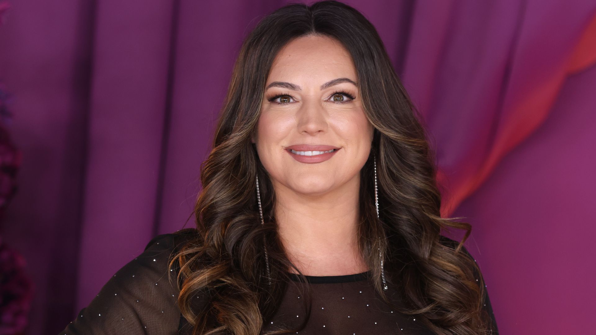 Kelly Brook smiles, wearing a sparkly black outfit and gold earrings