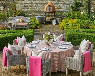 An example of garden picnic ideas showing an outdoor dining table with a red and white table cloth and pink blankets on the chairs in front of shrubbery and a BBQ area