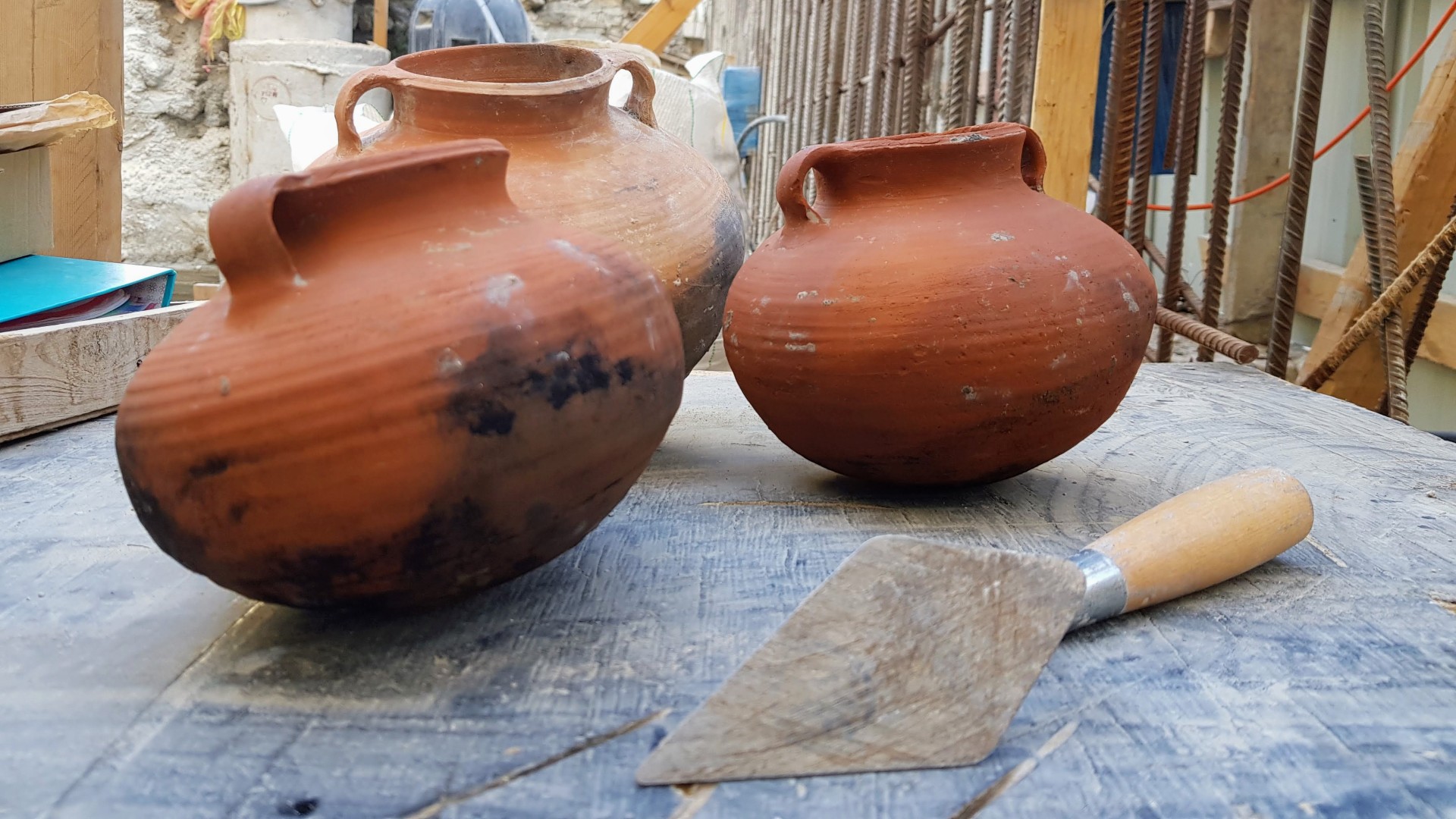 Three intact pottery cooking vessels. They have a bulbous shape with 2 small handles on either side at the top. There is a builder’s trowel in front of them.