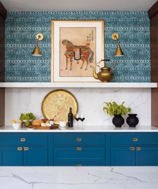A kitchen with a blue wallpapered wall with two gold wall sconces, dark blue cabinets with white countertops above with a cheese board and two black vases, and a marble kitchen island