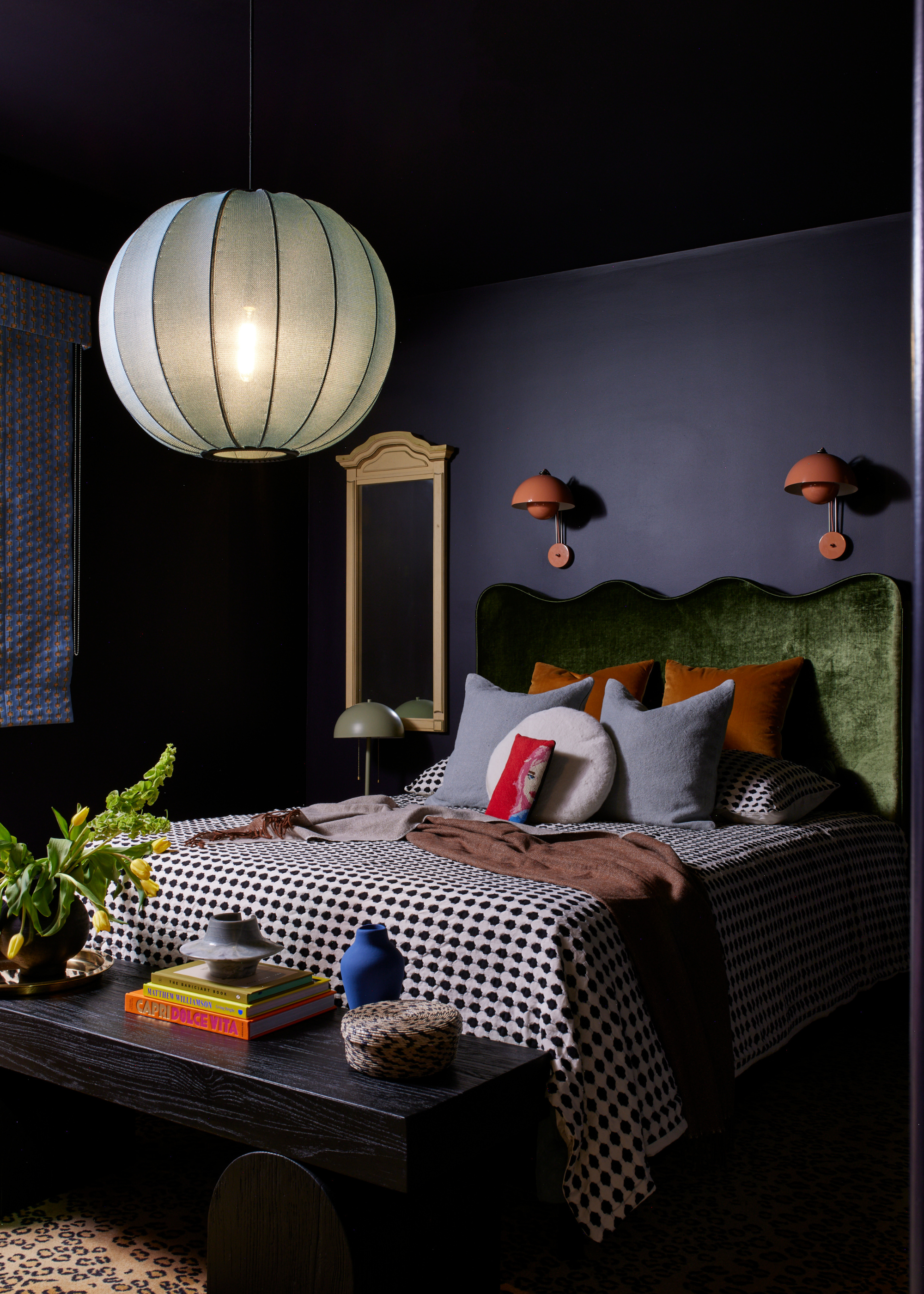 A dark blue bedroom with burnt orange wall sconces, an emarald green headboard, an abundance of pillows on a checkered bedscape, a semi-sheer lantern pendant, a black stool with books and vases