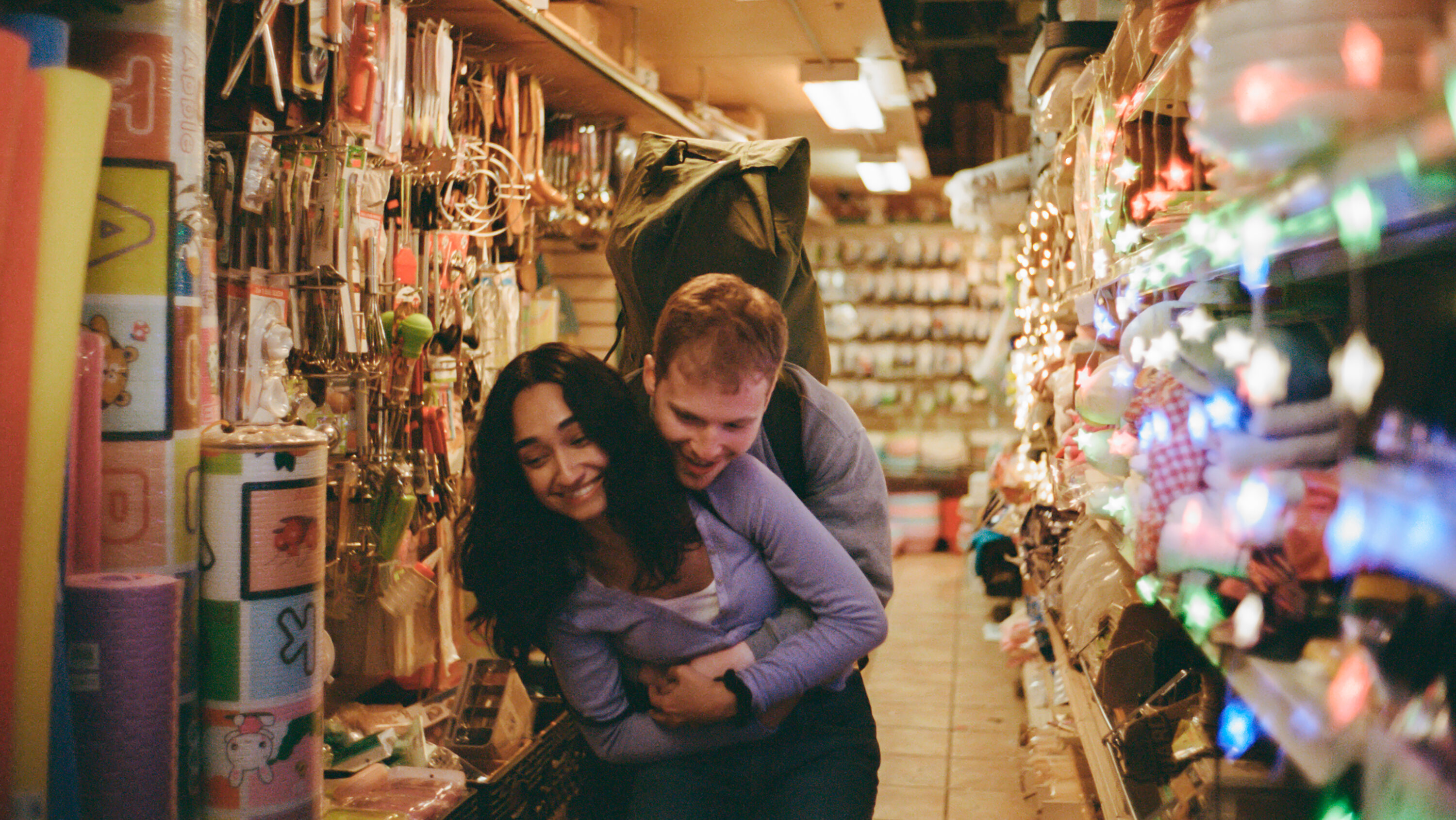 Fred Hechinger as Skinner hugging Sebiye Behtiyar as Aishe from behind in a busy gift shop in a still from the movie preparation for the next life