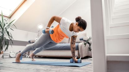 A woman in workout clothing performing a renegade row at home with dumbbells