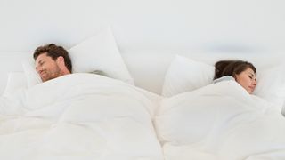 A couple sharing a bed and sleeping soundly. The man is sleeping on his back, and the woman is on her side, both are tucked up in a duvet