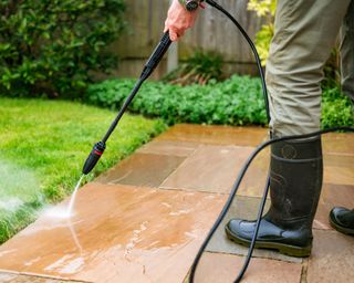 A senior man in his 70s cleans his patio stone slabs using a pressure washer. The man wears a green and blue check shirt with a green gilet