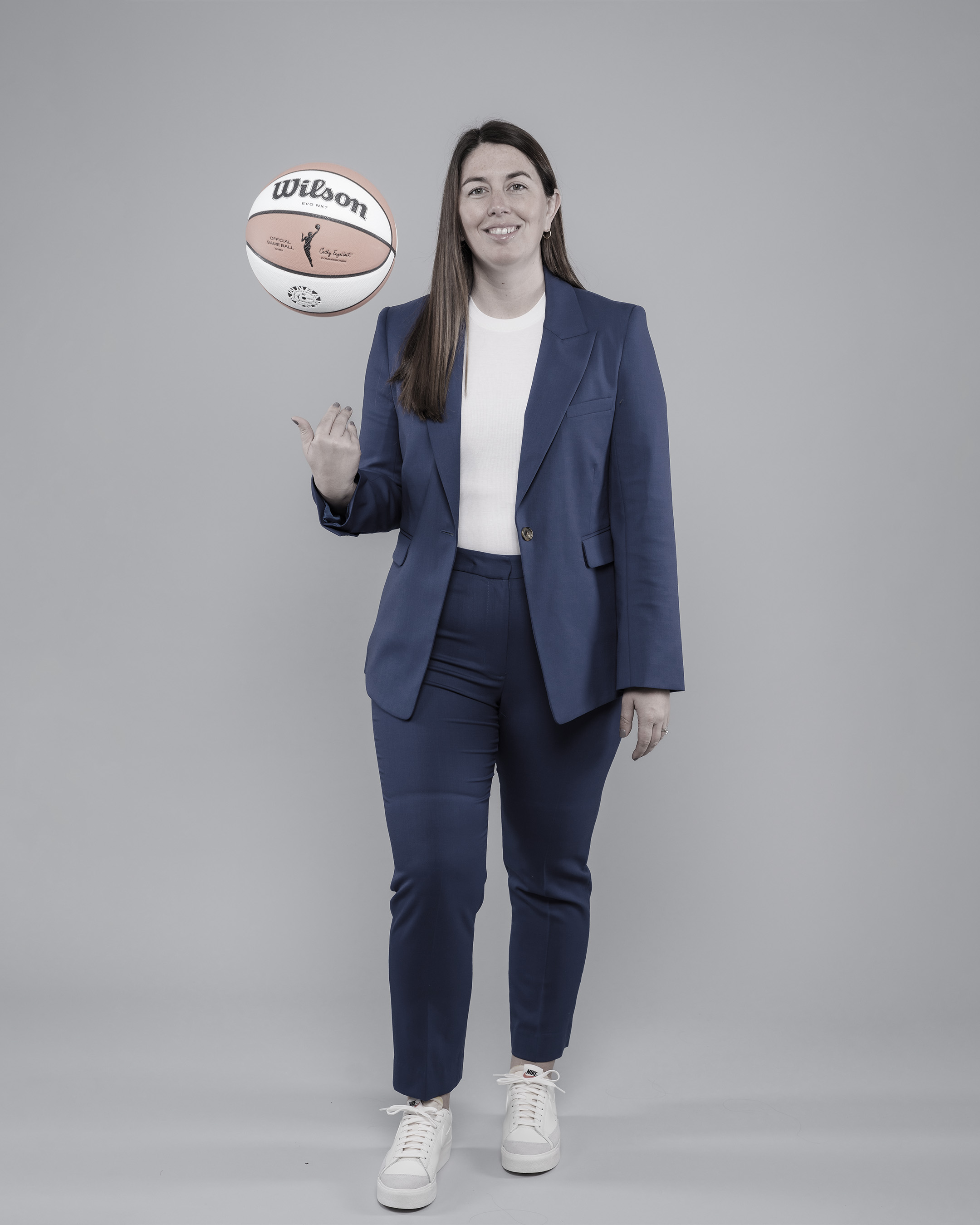Jess Smith standing in a blue suit, smiling, while tossing a basketball in the air.