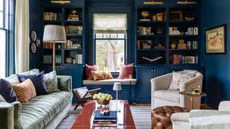 navy blue living room with custom bookcases built around a window with a nook seat, a velvet green couch, glossy coffee table and neutral tub chairs