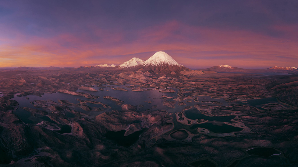 Aerial landscape of snow-capped volcanoes, rugged terrain, and interconnected lakes beneath a vivid purple-orange sunset