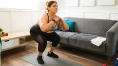 woman doing a squat on a grey yoga mat in a living room setting, with a grey sofa to the side of her. she's wearing black leggings, black trainers and a grey crop top.