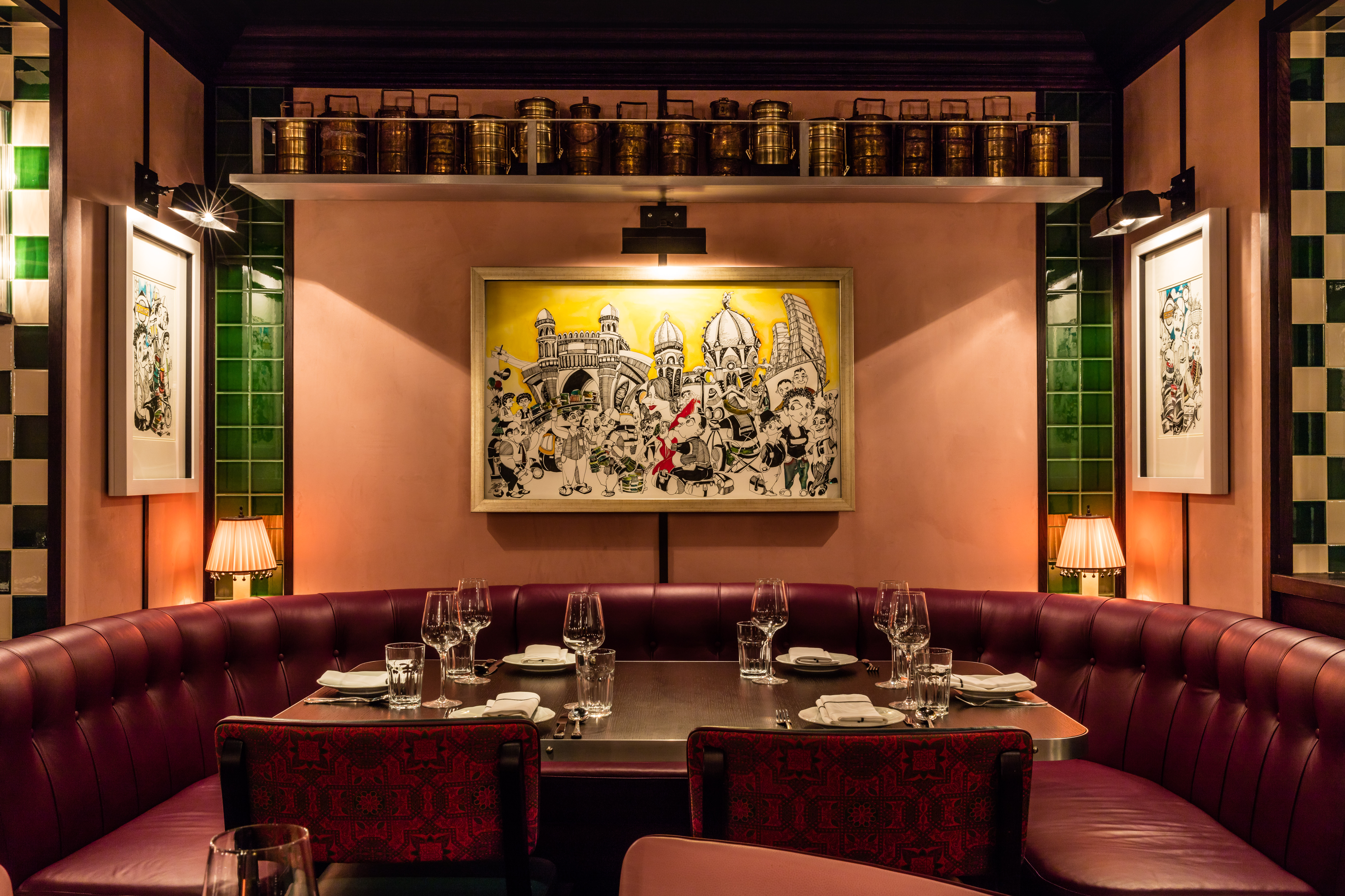 A colorfully decorated restaurant booth with red leather banquettes, green and white tiling, a framed artwork depicting a city's architectural skyline, and a series of jars resting on a shelf.