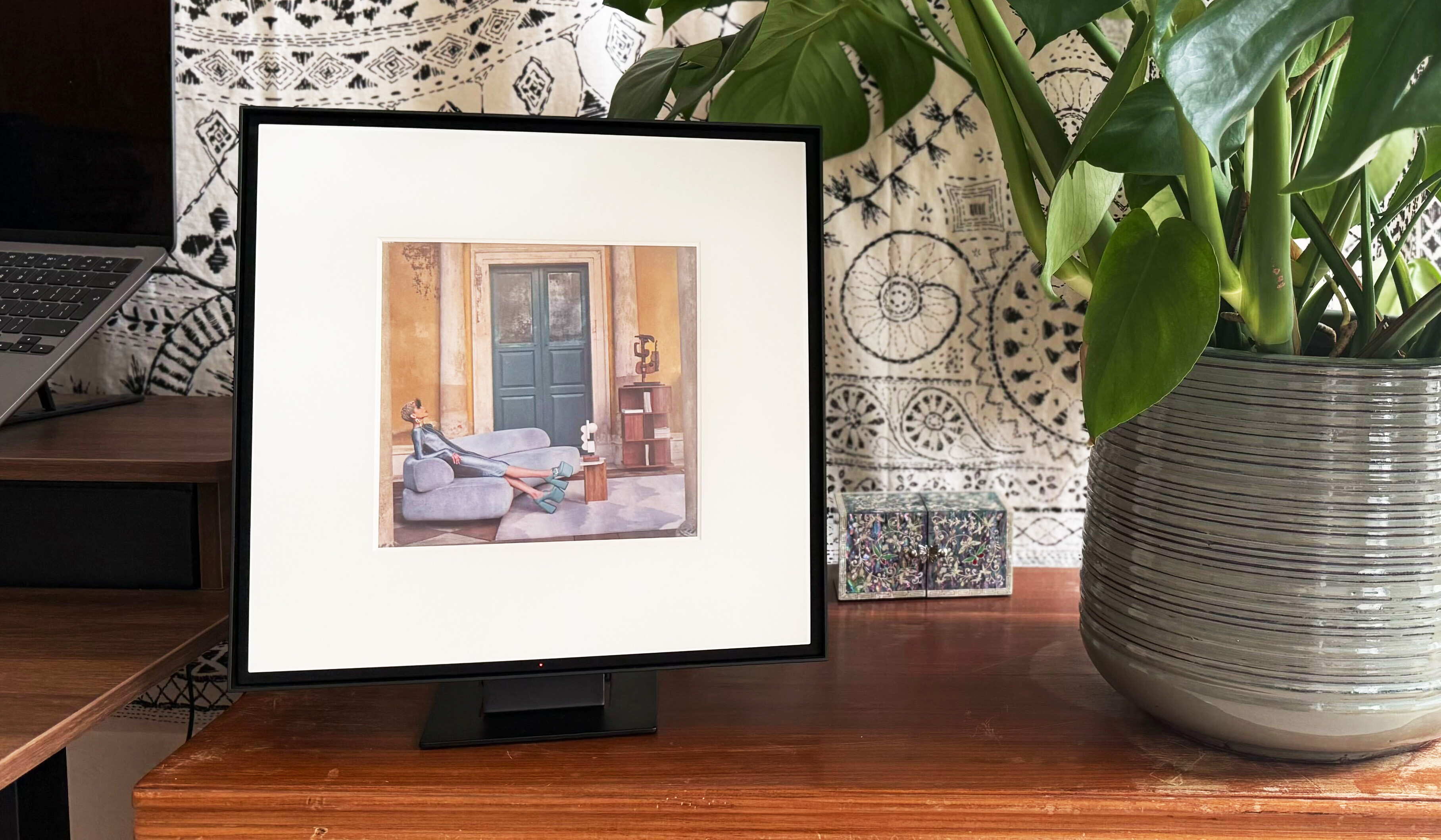 the samsung music frame speaker next to a monstera plant on a wood sideboard with a boho tapestry in the background