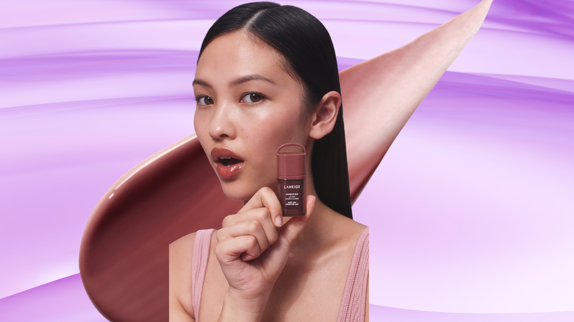 A model with black hair holding up a laneige lip tint. She is in front of an oversized swatch of the product, which is overlaid over a lavender, textured background. 