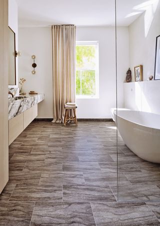 modern bathroom with white tub, glass shower wall, marble topped vanity, stool, cream drapes, vinyl floor