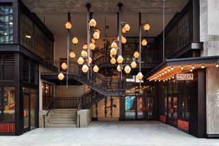 Daytime exterior image of the new Ace Hotel Brooklyn, stone entrance walkway with spiral staircase, black metal frame with red hand rails, glass walls seeing into internal ground floor area, multiple hanging orange diamond shaped lights above the walkway, hotel sign lit up and lights under canopy, upper level glass windows