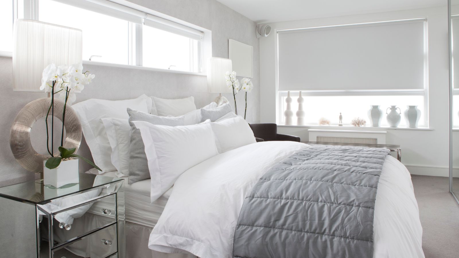 A white bedroom with large double bed with white bedding, mirrored chest of drawers, white blinds over a large window, and two long, large windows above the bed.