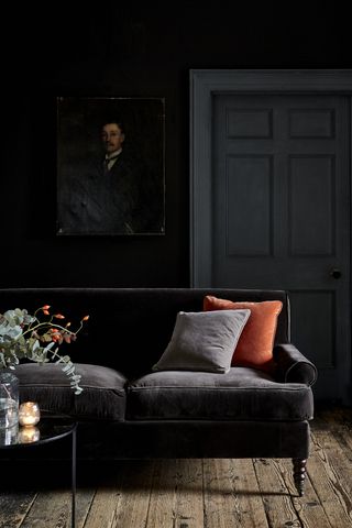 dark living room with black walls, wooden floors, grey door, chocolate brown couch, orange cushion, coffee table, artwork