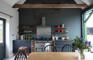 A dark grey kitchen with a background wall in the same color with open shelving, dining table and stainless steel cooking hood