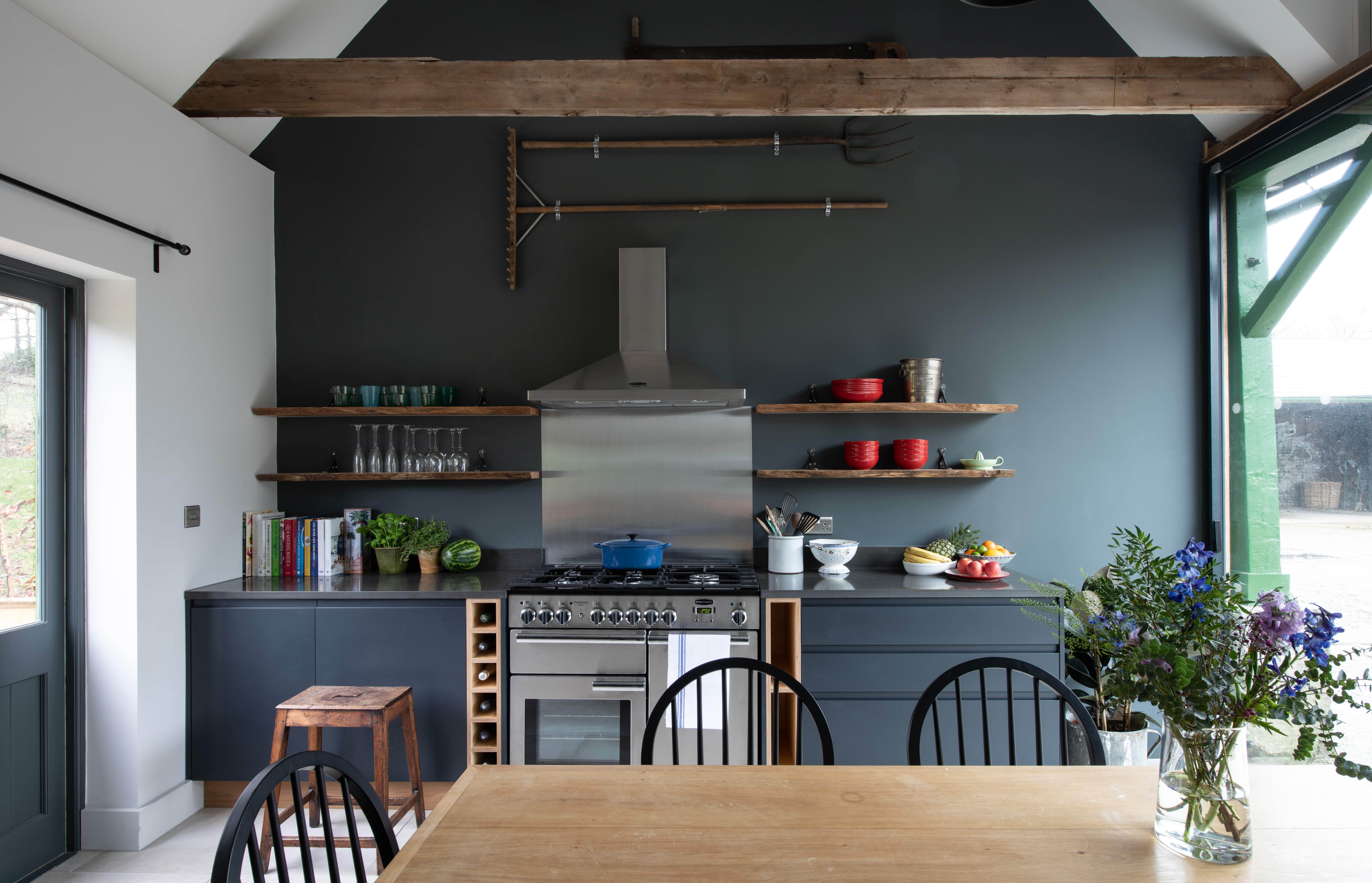 A dark grey kitchen with a background wall in the same color with open shelving, dining table and stainless steel cooking hood