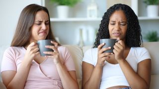 Two women sitting on sofa with cups of coffee, looking disgusted