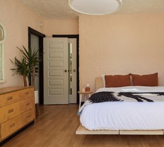 bedroom with low slung wooden bed, retro sideboard, pale pink/coral walls, pale green door and mirror, black woodwork, russet bed pillows, side table, plants