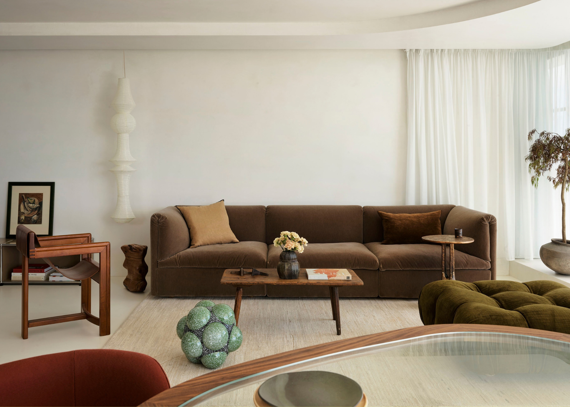 A white living room with an architectural pendant, a brown sofa, a green sofa, a turquoise statue, a wood coffee table, and a sling chair