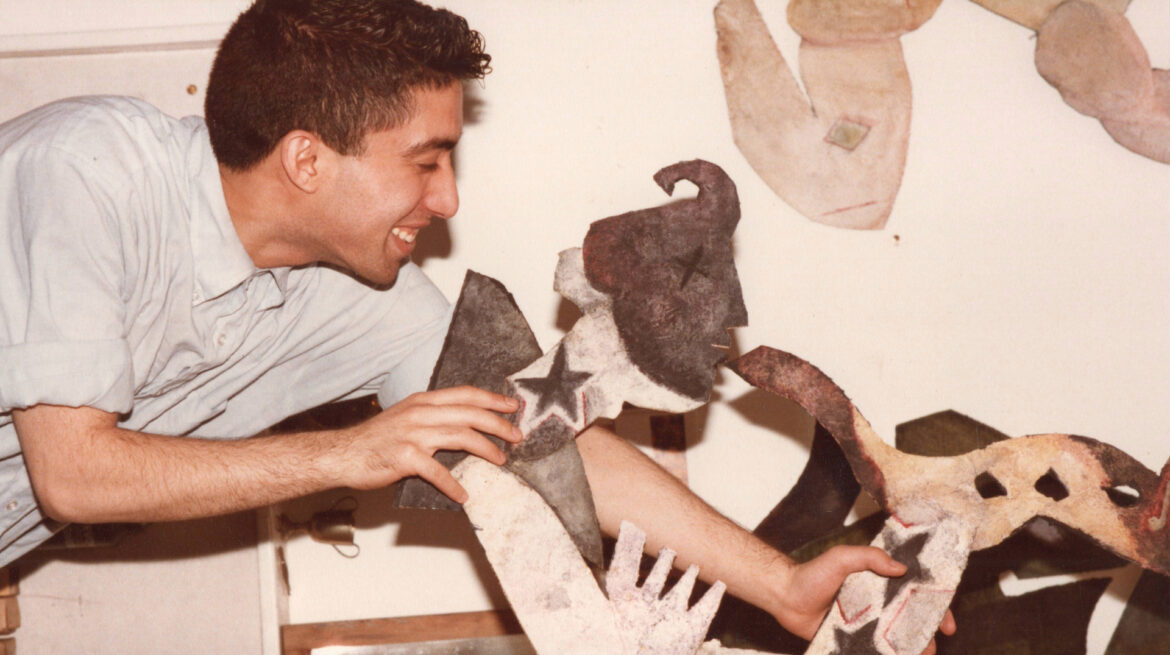 A young man dressed in a light blue shirt with folded-up sleeves plays with some anthopomorphic paper sculptures in a warmly lit room.