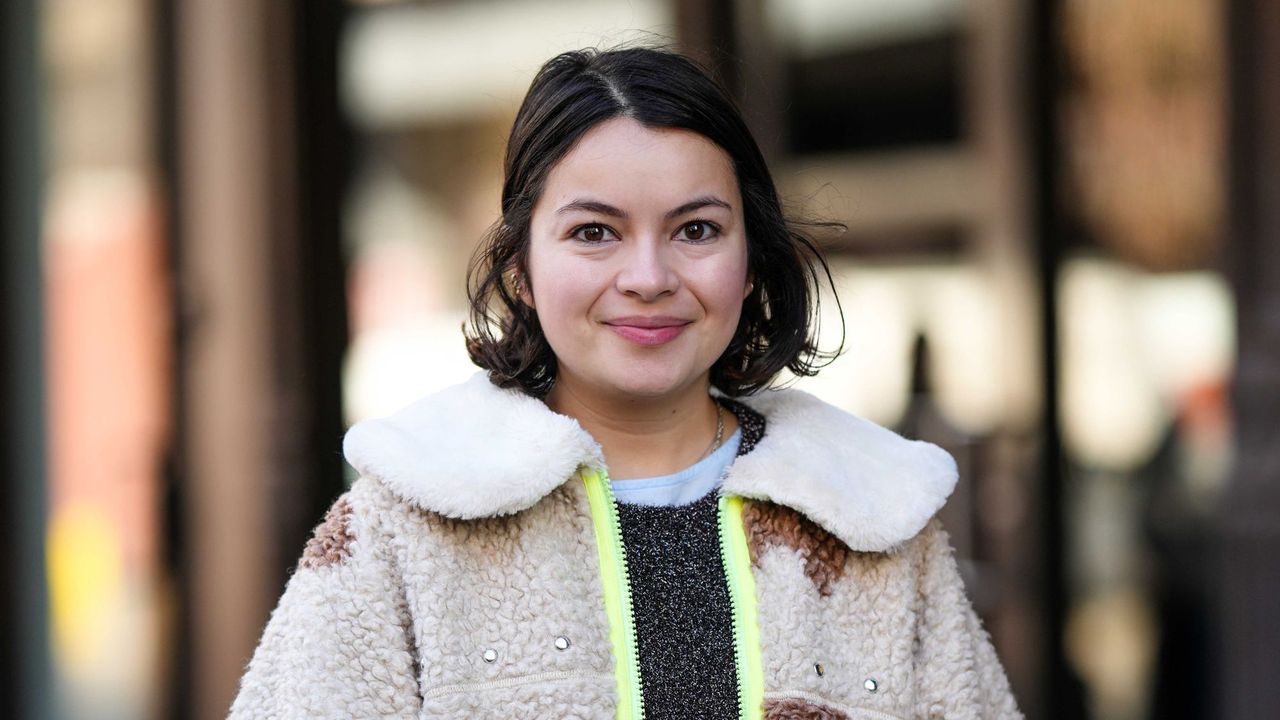 A smiley girl wears a pale blue long ruffled dress, a dark gray wool pullover, a beige and brown print pattern sheep zipper jacket