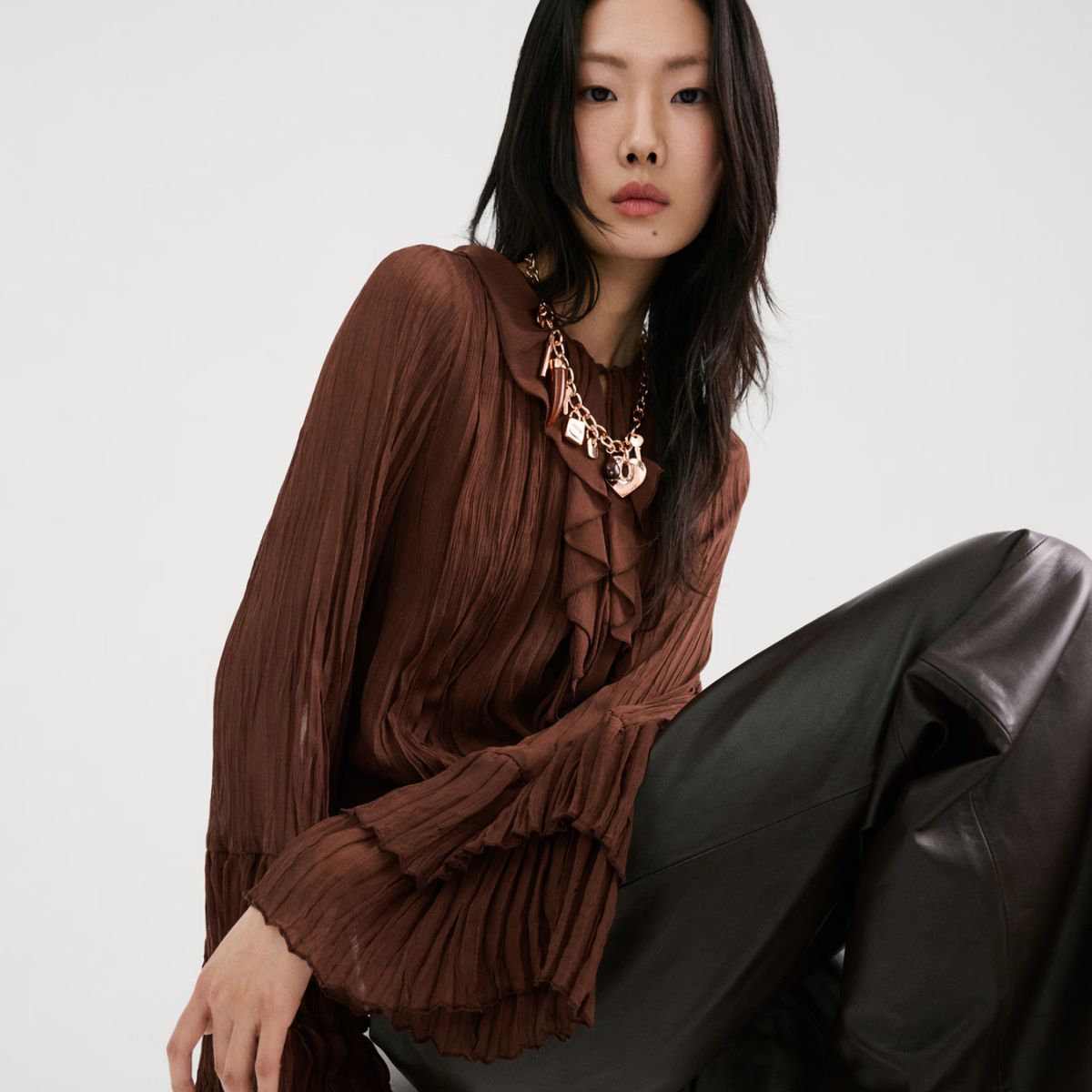 Model wears brown pleated blouse, gold charm necklace, leather trousers