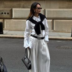 Caroline wears a white shirt, white trousers, jumper over shoulders and carries black handbag
