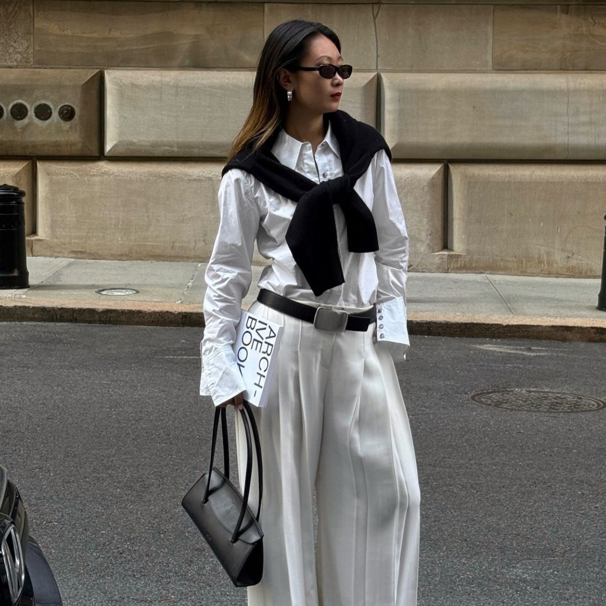 Caroline wears a white shirt, white trousers, jumper over shoulders and carries black handbag