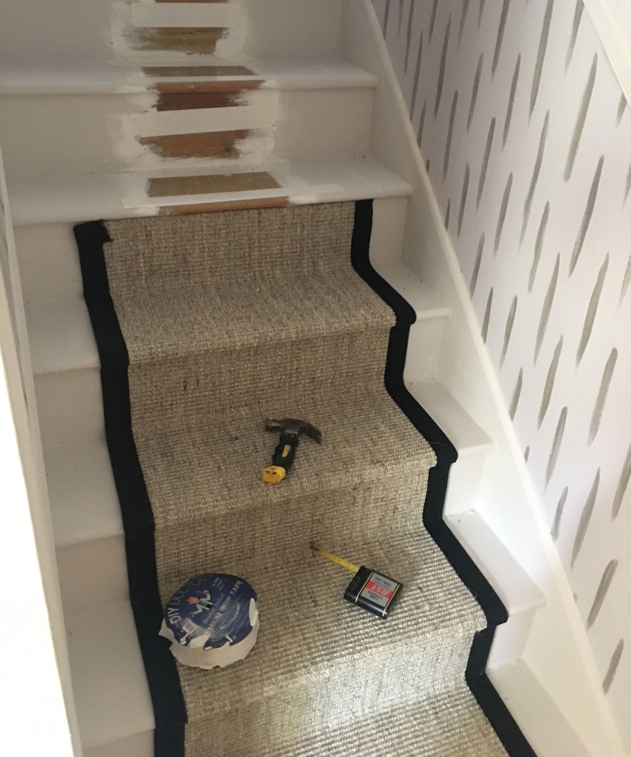 A beige woven stair runner with black edging being installed on a white staircase with a hammer, tape and tape measure on it