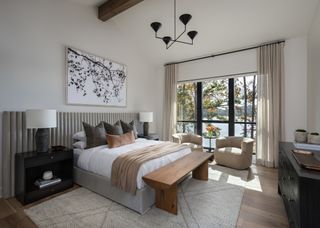 A modern bedroom with a neutral color palette, a wooden bench at the end of the bed, and two accent chairs in the window