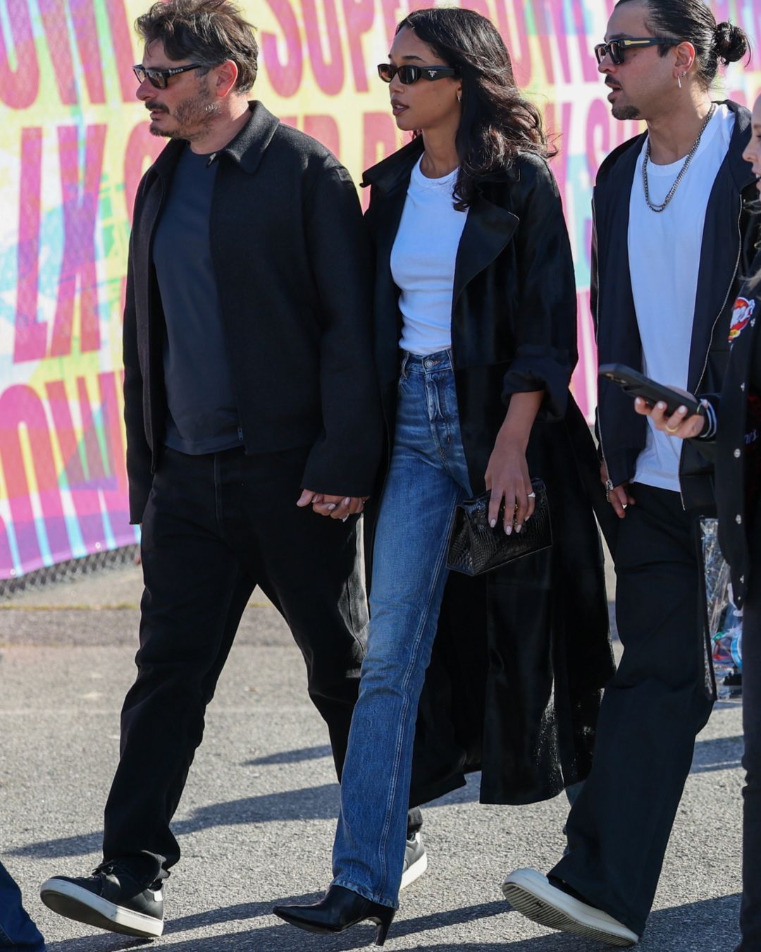 Laura Harrier walks outside wearing high-waisted straight-leg jeans with heeled boots, a white tee and a black trench coat.