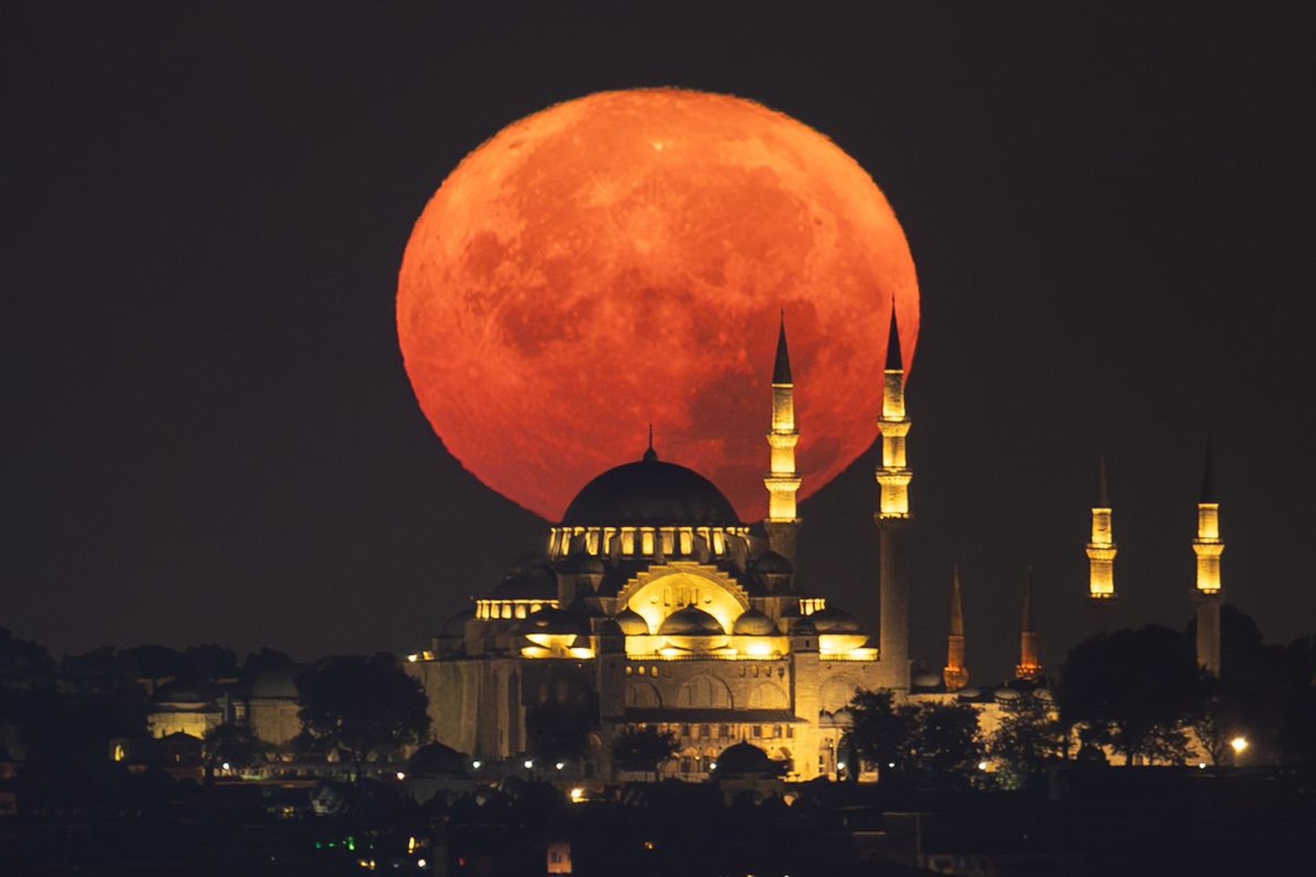 A massive orange moon rising behind a lit-up mosque