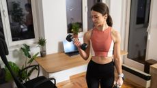 woman in a home setting holding a dumbbell in each hand, curling one up toward her shoulder and looking at it. she's wearing a coral crop top and black leggings. 