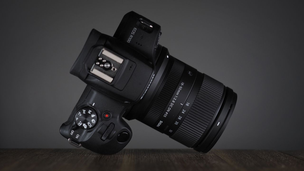 The Sigma 18-50mm f/2.8 DC DN | C Canon RF lens, mounted to a Canon EOS R100, on a wooden surface against a dark background with moody lighting