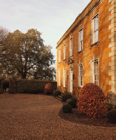 a stone 1700s English country home with a shingle driveway and box hedges