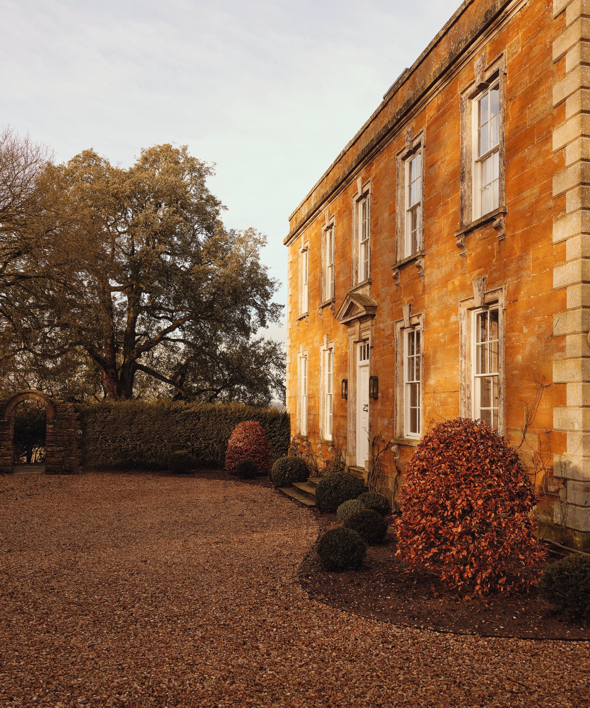 a stone 1700s English country home with a shingle driveway and box hedges