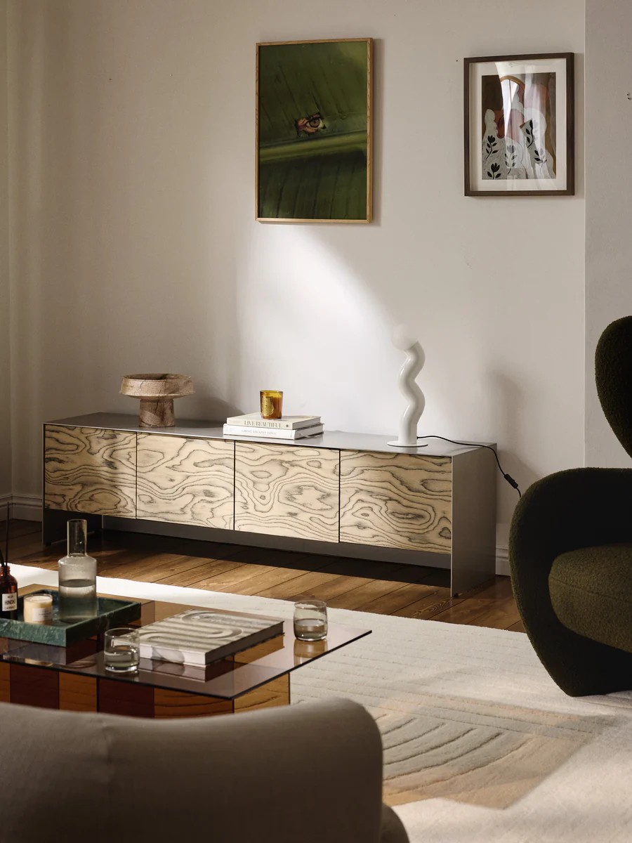 Image of a long and low sideboard with black and white wood grain on the doors and a brushed chrome frame. It's styled in a contemporary room with lots of lamps, vases, and coffee table books.