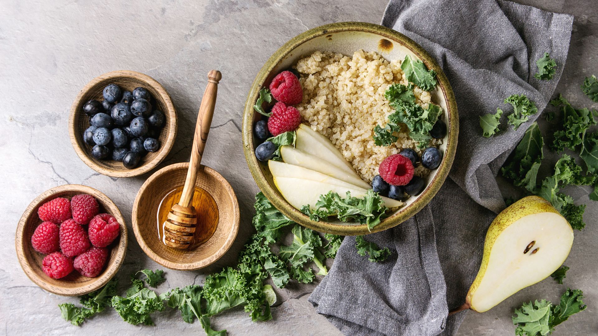 Vegan quinoa porridge with kale, strawberries, blueberries, sliced pear, honey on dipper in bowl with ingredients above and cloth napkin over grey texture background Top view,