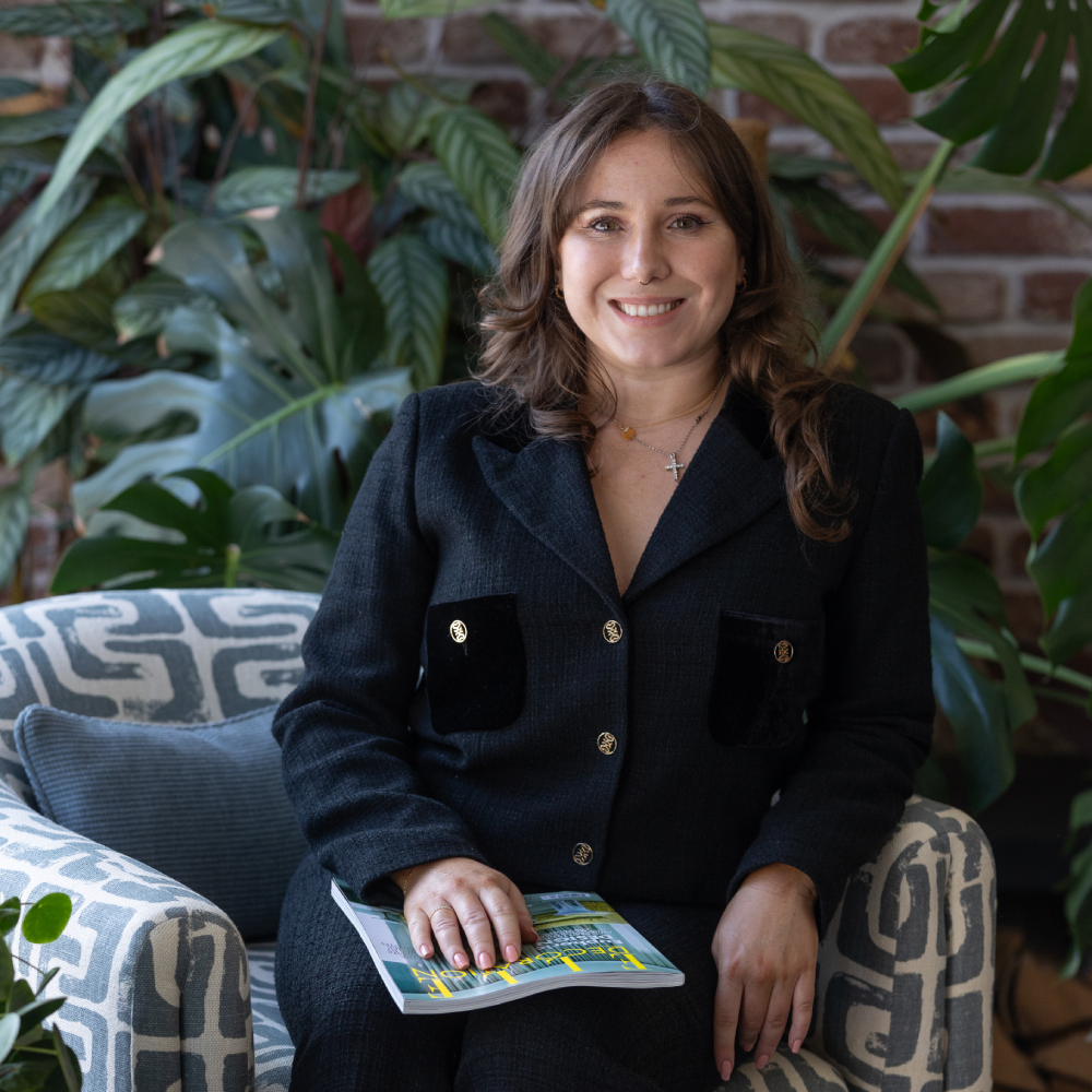 female with long brown hair wearing black clothing sat on patterned armchair with plants in background