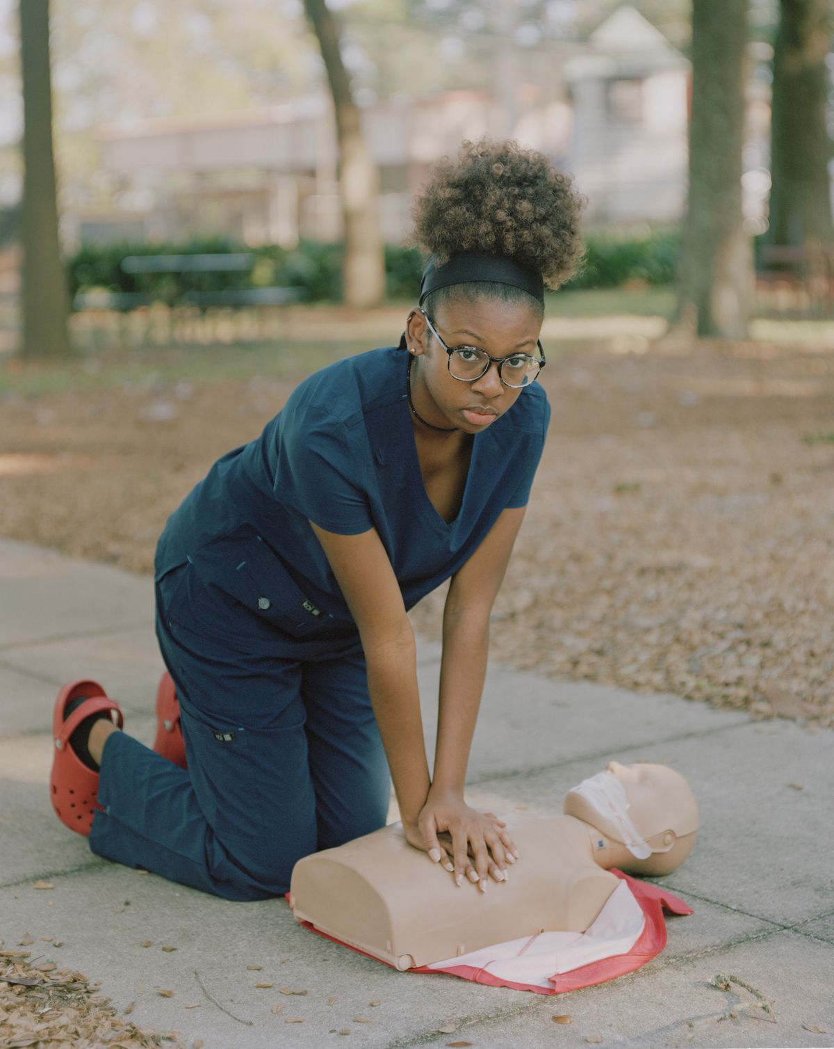 A girl in navy scrubs and red clogs kneels on a park path, practising chest compressions on a CPR training dummy