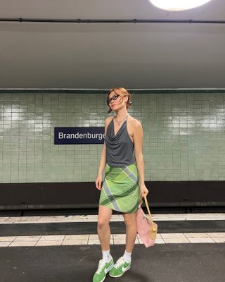 Vivian parada en una estación de metro con una blusa sin mangas gris y una falda a cuadros verde con zapatillas verdes