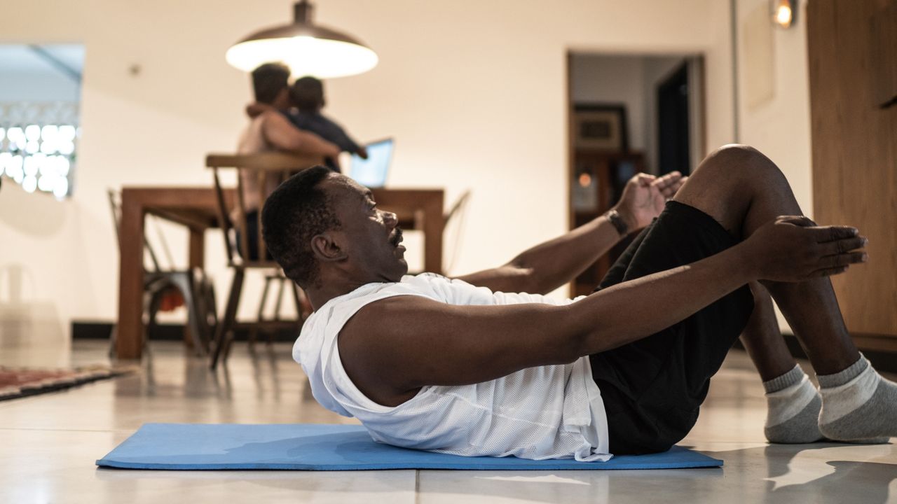 man wearing white vest and black shorts doing a pilates roll up movement on a blue mat in a home setting. There&#039;s a dining table with a female and child sat on a chair in the background.