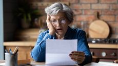 An older woman looks dismayed as she looks at paperwork while sitting at a table in her kitchen.