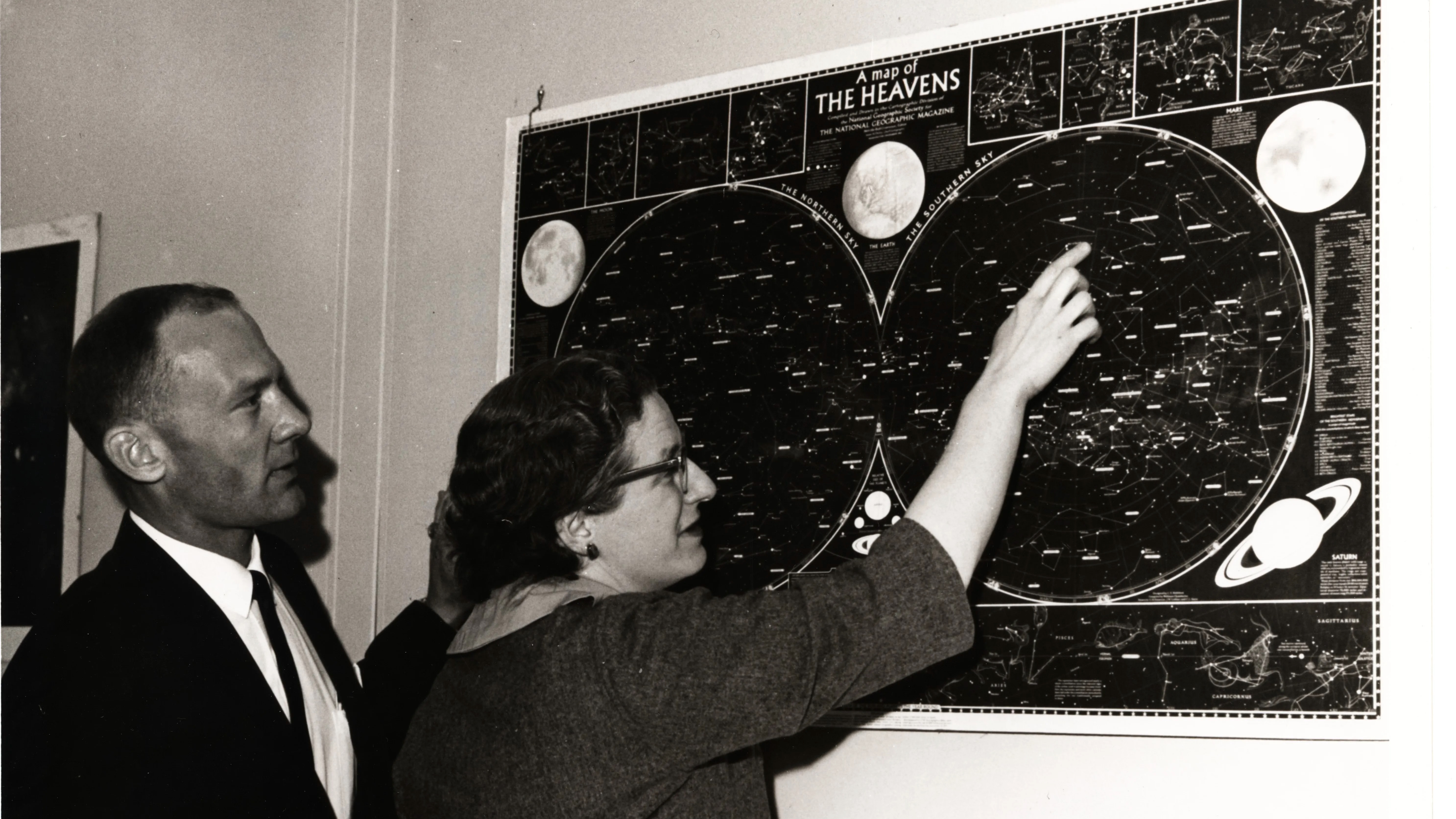black and white photo of a woman showing a man a wall-mounted star chart