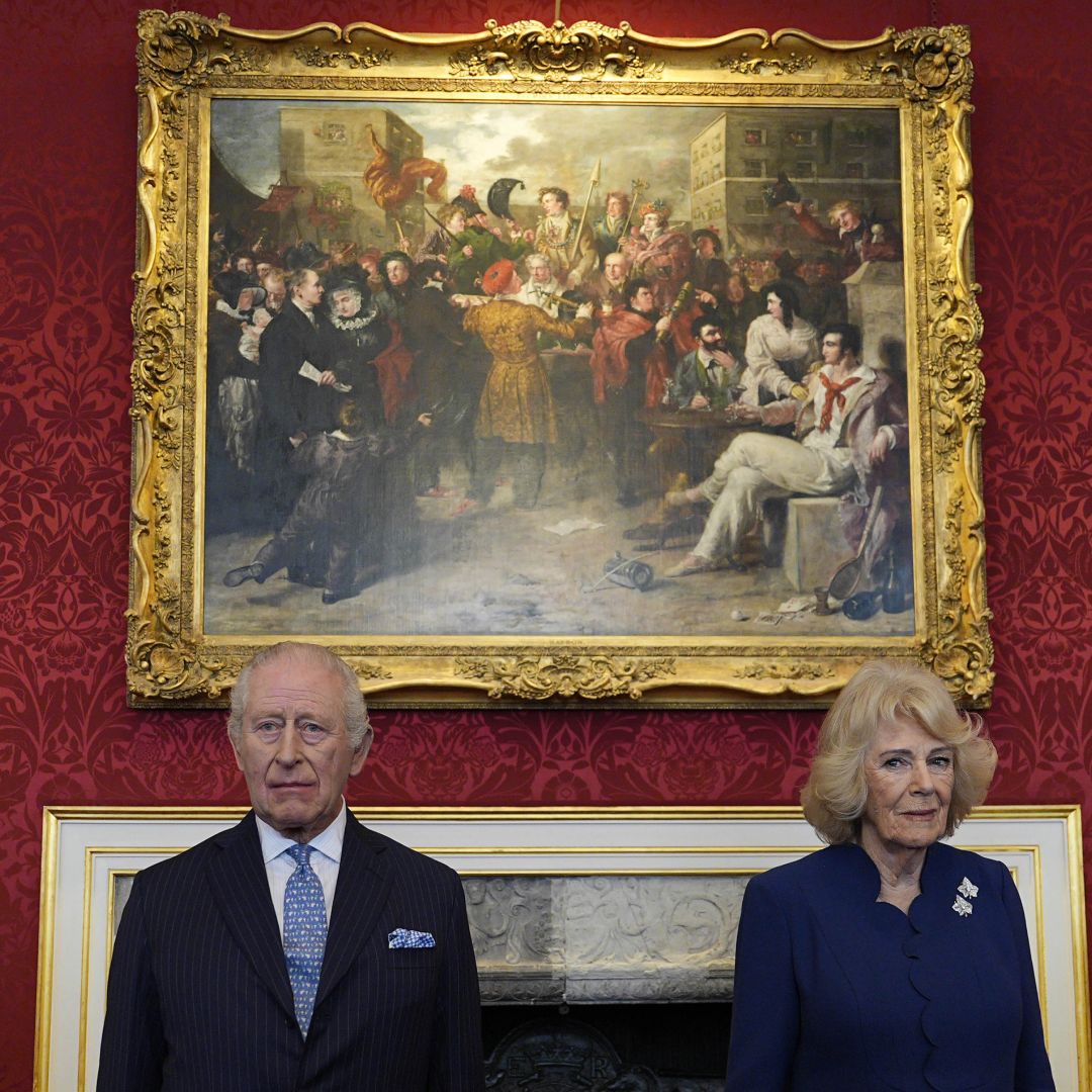 King Charles and Queen Camilla standing in front of a painting on a red wall with serious expressions