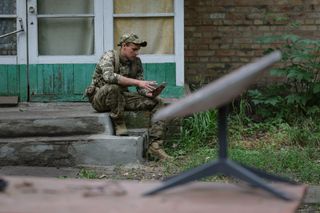 a soldier in camouflage sits on a porch looking at a cell phone behind a small portable satellite dish