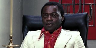 Yaphet Kotto in a white suit with red shirt sitting behind a desk.