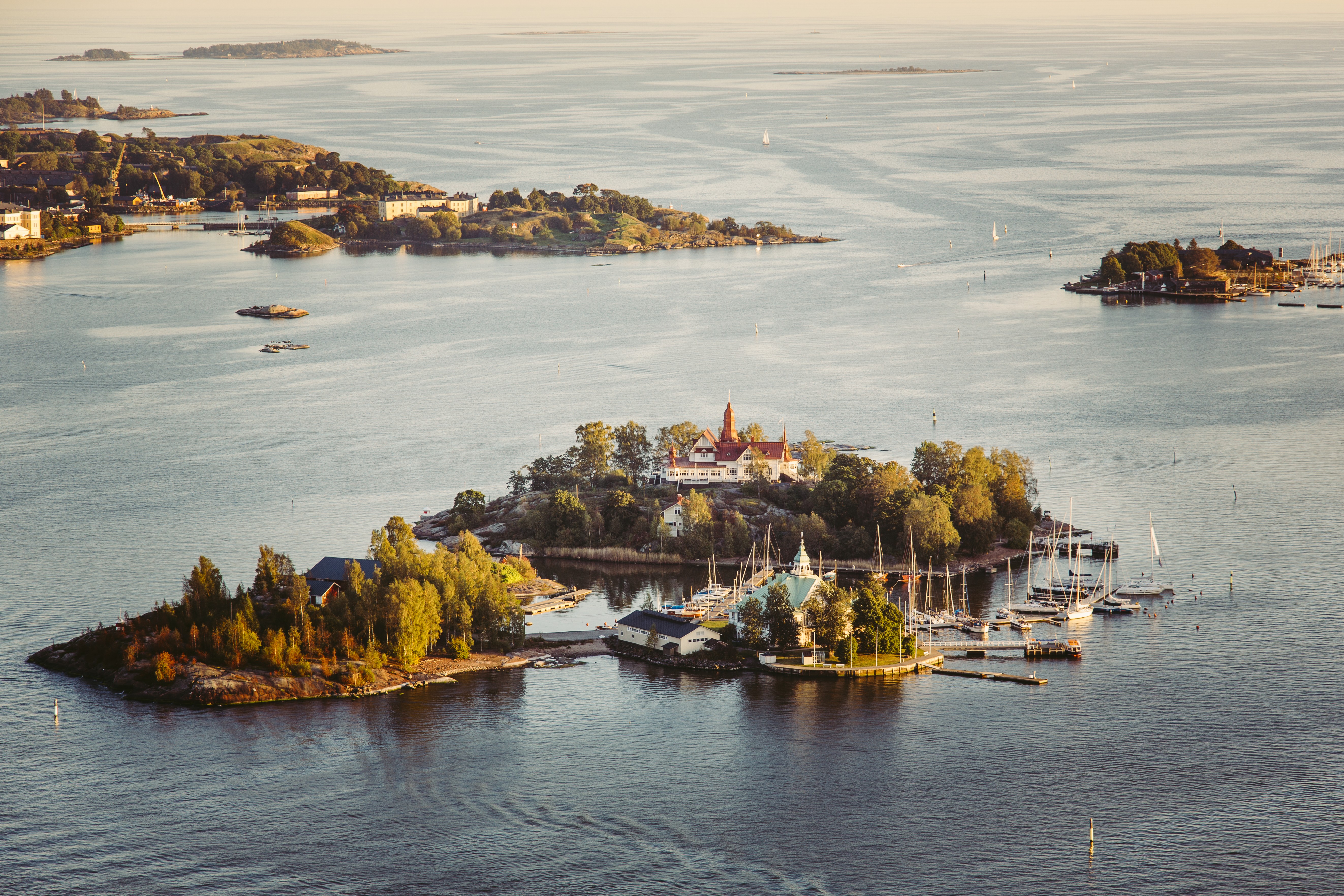 A sun-lit island with just a few buildings, lots of sailing boats, and dense vegetation is photographed from high above on a sunny day.